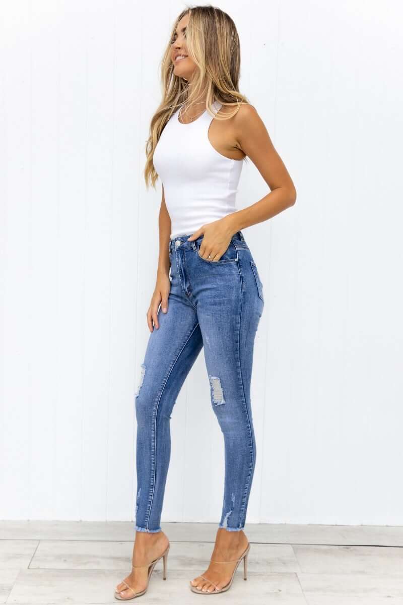 Model wearing Qilli distressed denim jeans with pockets and raw hem, styled with a white tank top and high heels.