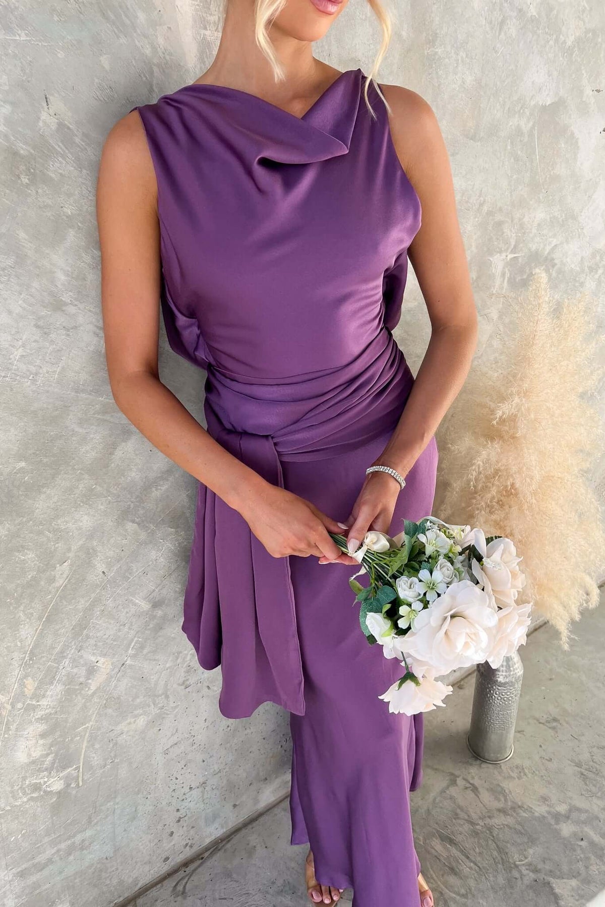 Qalenna Maxi Dress in purple satin, high neckline, featuring open back and styled with bouquet of flowers.