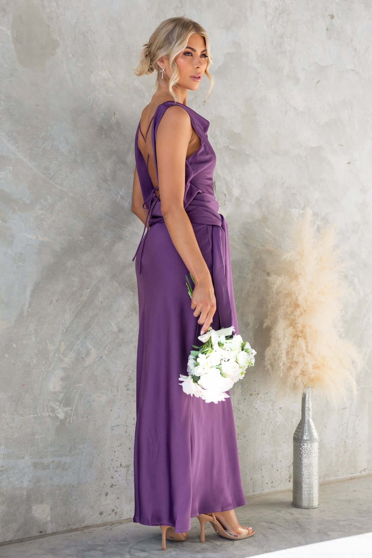 Woman in a purple Qalenna maxi dress with open back and tie, holding white flowers against a textured wall.
