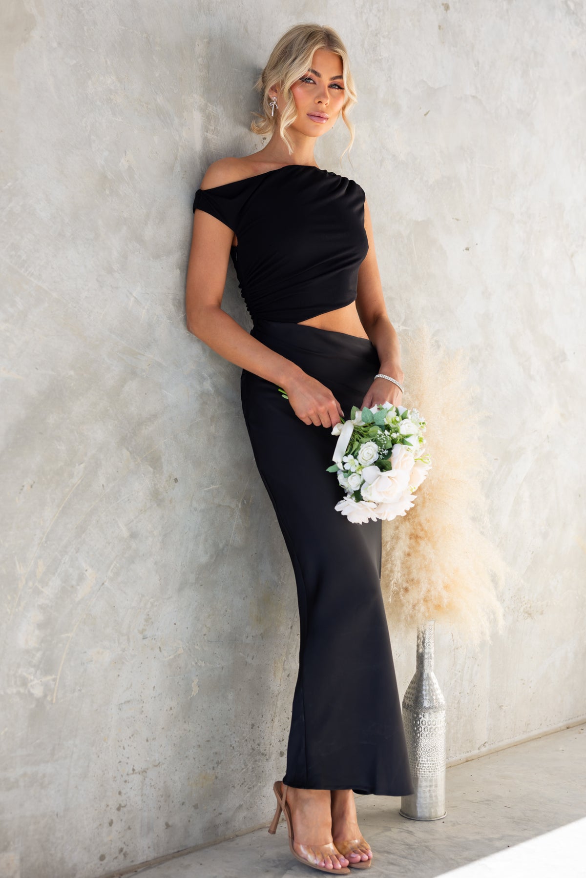 Model wearing Queta Maxi Dress with asymmetric neckline and satin skirt, holding a floral bouquet against a textured wall.