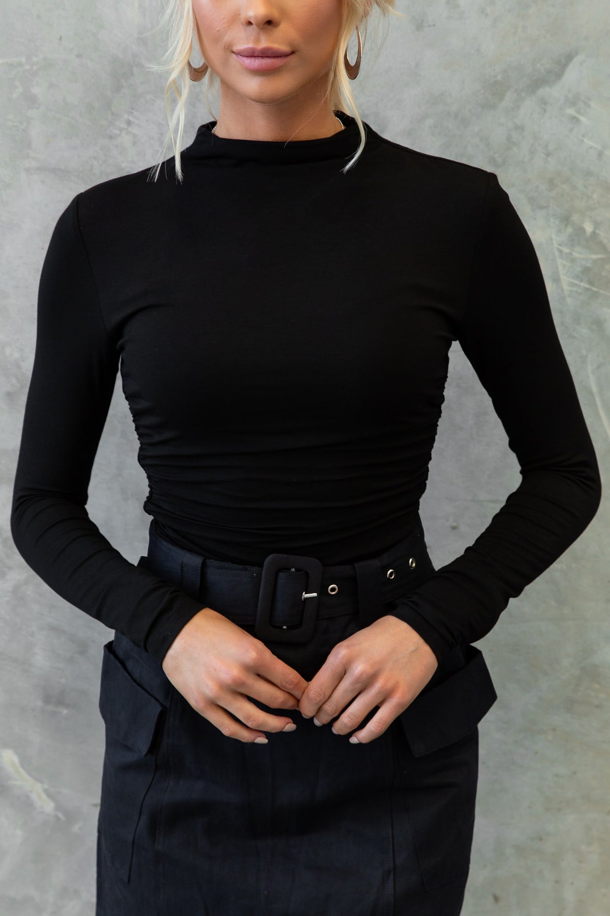 Model wearing Rumba Top, a black high-necklined top with a fitted design, paired with a belted skirt.