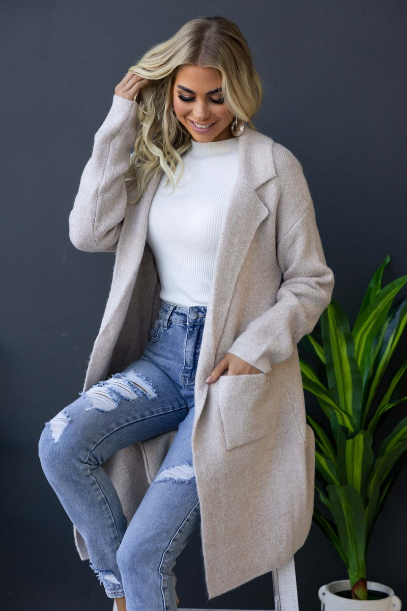 Sarlia Cardigan in light beige with pockets, worn over a white top and distressed jeans, styled with indoor plant backdrop.